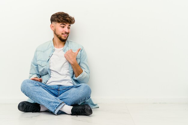 Jonge Marokkaanse man zittend op de vloer wijst met duimvinger weg, lachend en zorgeloos.