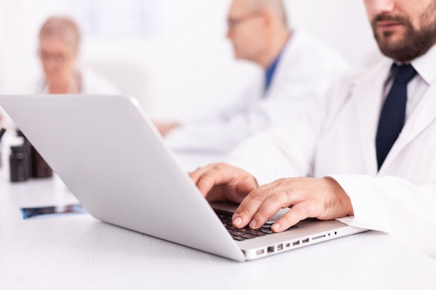 Jonge medisch specialist typen op laptop tijdens ontmoeting met collega's in ziekenhuiskantoor. Kliniekdeskundige therapeut in gesprek met collega's over ziekte, medisch professional