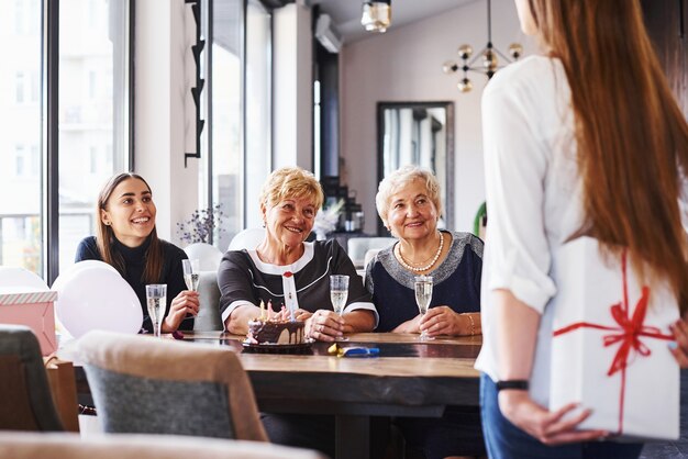 Jonge meisjes houden geschenkdozen achter zich. Senior vrouw met familie en vrienden die een verjaardag binnenshuis viert.