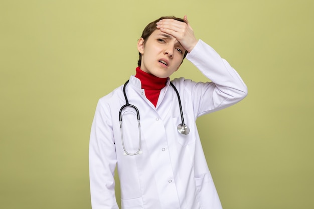 Jonge meisjesdokter in witte jas met stethoscoop om nek die verward en erg angstig kijkt met de hand op haar voorhoofd voor een fout die over de groene muur staat