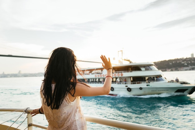 Jonge meisjesreiziger ontspant tijdens een cruise op een jacht, zwaaiend met haar hand naar een passerend schip bij zonsondergang, een brug tegen een achtergrond