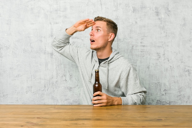 Jonge mens die een bier op een lijst drinkt die ver weg houdend hand op voorhoofd kijkt.