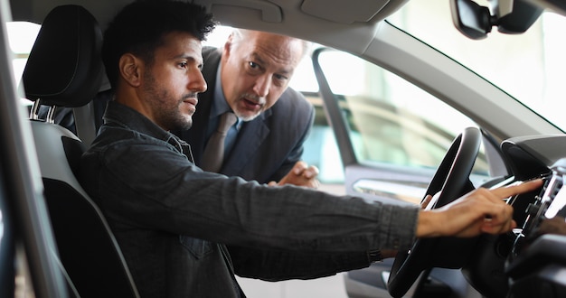 Jonge mens die een nieuwe auto in een toonzaal onderzoekt terwijl de autohandelaar zijn eigenschappen verklaart