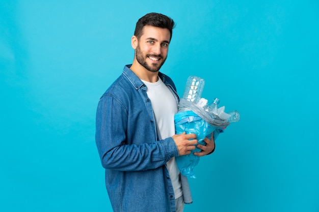 Jonge mens die een zakhoogtepunt van plastic te recycleren flessen houden geïsoleerd op blauwe muur die veel glimlachen