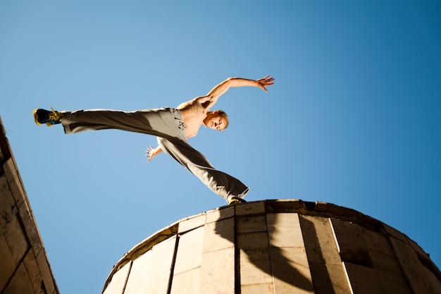 Jonge mens die en parkour tussen gebouwendaken buiten springen oefenen op duidelijke de zomerdag met blauwe hemel bij achtergrond