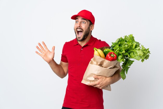 Jonge mens die met baard een zakhoogtepunt van groenten houdt die op witte muur met verrassingsgelaatsuitdrukking wordt geïsoleerd