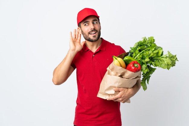 Jonge mens die met baard een zakhoogtepunt van groenten houdt die op witte muur wordt geïsoleerd luisterend iets