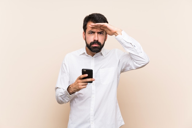 Jonge mens die met baard mobiel houden kijkend ver weg met hand om iets te kijken