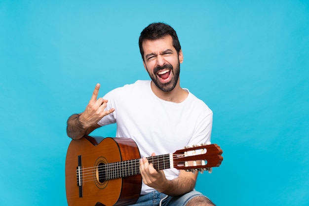 Jonge mens die met gitaar over geïsoleerde blauwe muur rotsgebaar maakt
