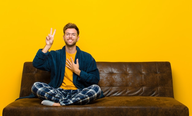 Jonge mens die pyjama's draagt die gelukkig, zeker en betrouwbaar kijken, en overwinningsteken, met een positieve houding glimlachen tonen. zittend op een bank