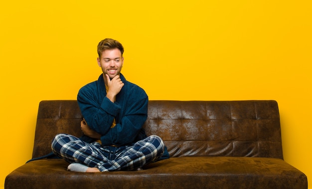 Jonge mens die pyjama's draagt die met een gelukkige, zekere uitdrukking met hand op kin glimlachen, en aan de kant benieuwd zijn kijken. zittend op een bank