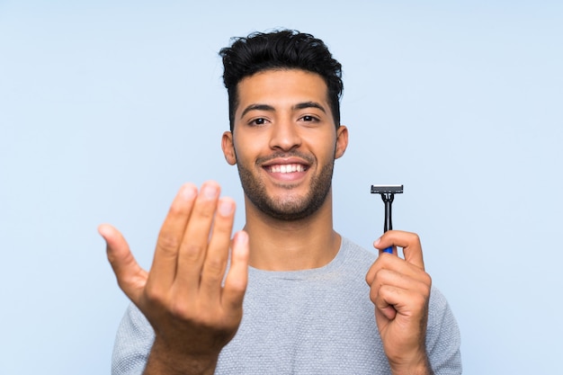 Jonge mens die zijn baard over geïsoleerde blauwe muur scheert die met hand uitnodigt te komen. Blij dat je bent gekomen