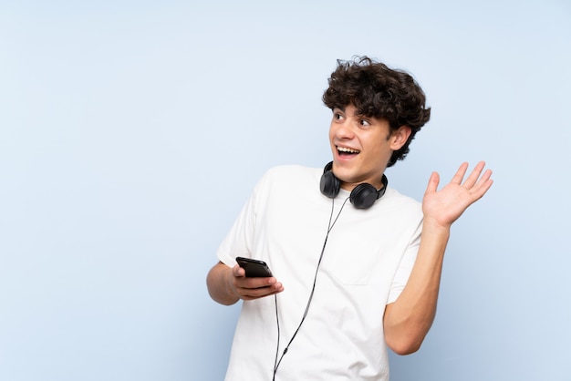 Jonge mens het luisteren muziek met mobiel over geïsoleerde blauwe muur met verrassingsgelaatsuitdrukking