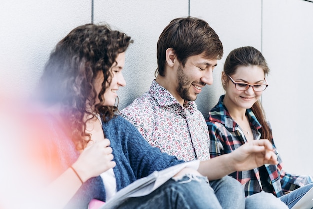 Jonge mensen gebruiken verschillende gadgets en glimlachen, zitten in de buurt van de muur. Studenten studeren met behulp van een laptop computer. Onderwijs social media concept.