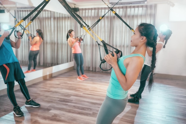 jonge mensen trainen binnenshuis met TRX die keihard trekt vol kracht. aziatische studenten in gym training armen sterkere borst. groep gelukkige vrienden die samen met riemen trainen.