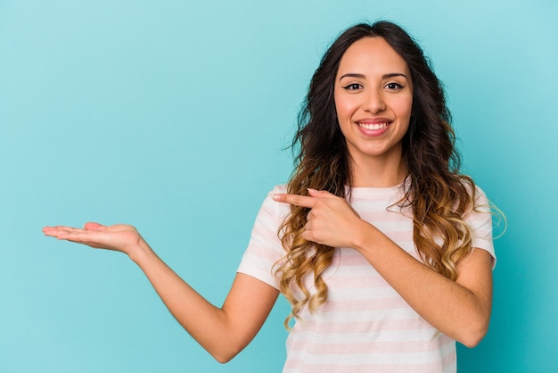 Jonge Mexicaanse vrouw geïsoleerd op blauwe muur opgewonden met een kopie ruimte op palm.