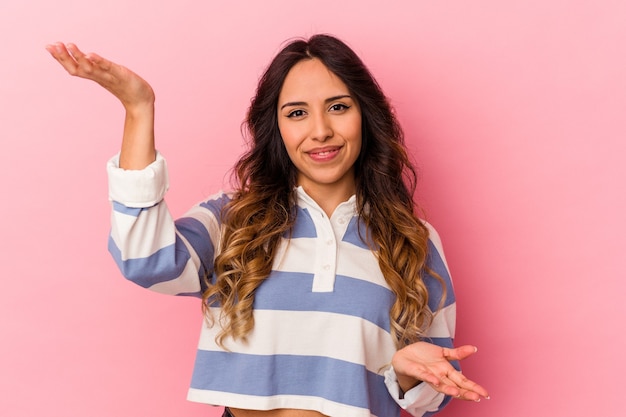 Jonge mexicaanse vrouw geïsoleerd op roze achtergrond maakt schaal met armen, voelt zich gelukkig en zelfverzekerd.