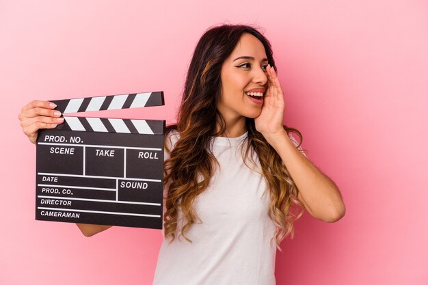 Jonge Mexicaanse vrouw met Filmklapper geïsoleerd op roze achtergrond schreeuwen en houden palm in de buurt van geopende mond.