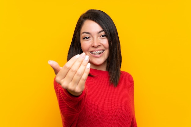 Jonge Mexicaanse vrouw met rode sweater over gele muur die uitnodigt te komen