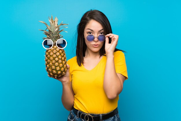 Foto jonge mexicaanse vrouw over geïsoleerd blauw die een ananas met zonnebril houdt