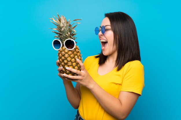 Jonge Mexicaanse vrouw over geïsoleerde blauw met een ananas met zonnebril