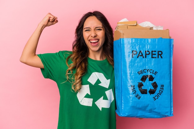 Jonge Mexicaanse vrouw recycling karton geïsoleerd op roze vuist verhogen na een overwinning, winnaar concept.