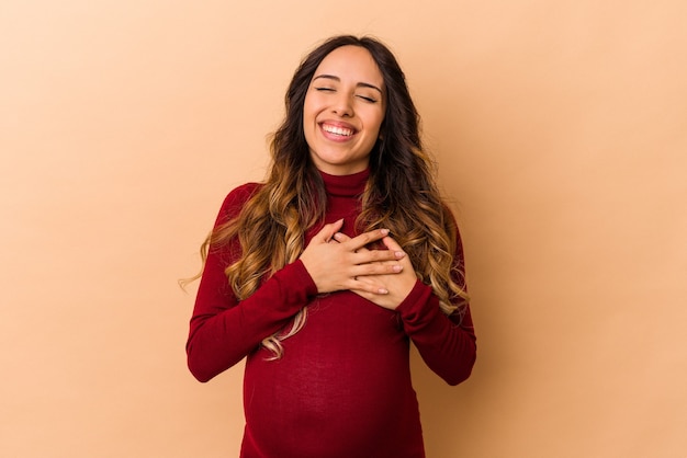 Jonge Mexicaanse zwangere vrouw geïsoleerd op beige achtergrond lachen hand in hand op het hart, concept van geluk.