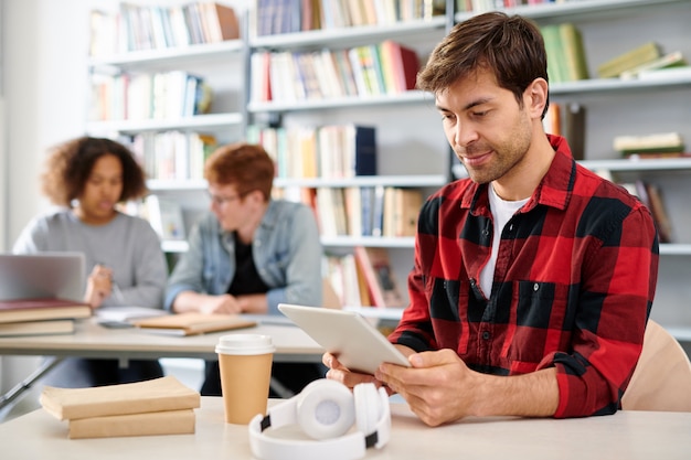 Jonge mobiele student met touchpad kijken naar online video door bureau tijdens pauze tussen lessen