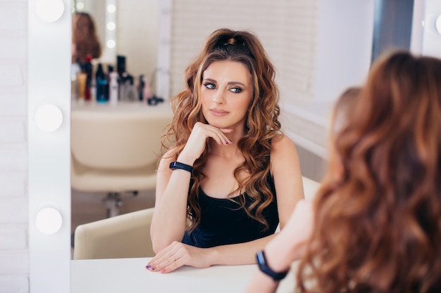 Jonge mode vrouw met brunette kapsel en make-up kijken naar de spiegel in de kapsalon