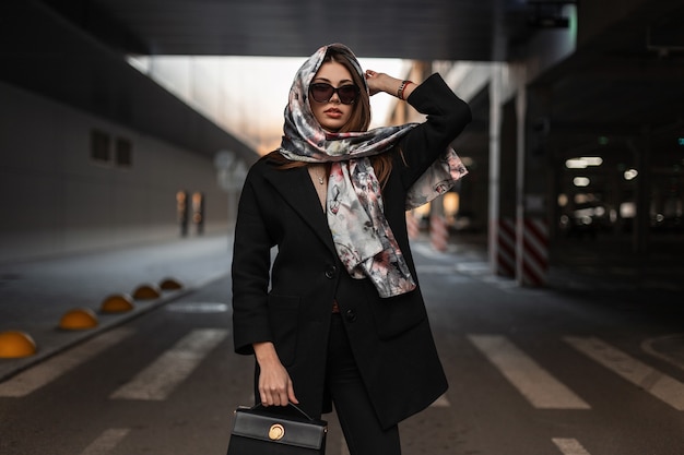 Jonge mode zakenvrouw in stijlvolle zonnebril in een zwarte jas met een modieuze lederen handtas in glamour sjaal poseren in de buurt van parkeerplaats op straat. Aantrekkelijk trendy meisje. Elegante dame.