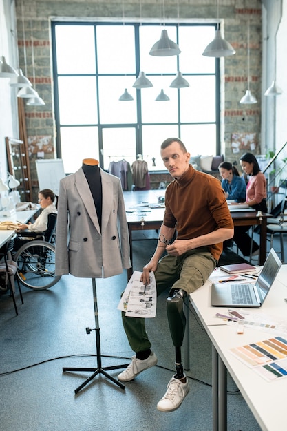 Jonge modeontwerper uitschakelen met schetsen zittend op tafel voor camera tegen drukke vrouwelijke collega's in grote werkplaats