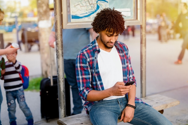 Jonge moderne Afro-Amerikaanse man zittend op een busstation en het gebruik van telefoon tijdens het wachten op zijn transport.