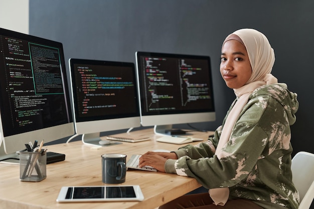 Jonge moderne it-specialist in hijab en sweatshirt die informatie op computerschermen decodeert terwijl si