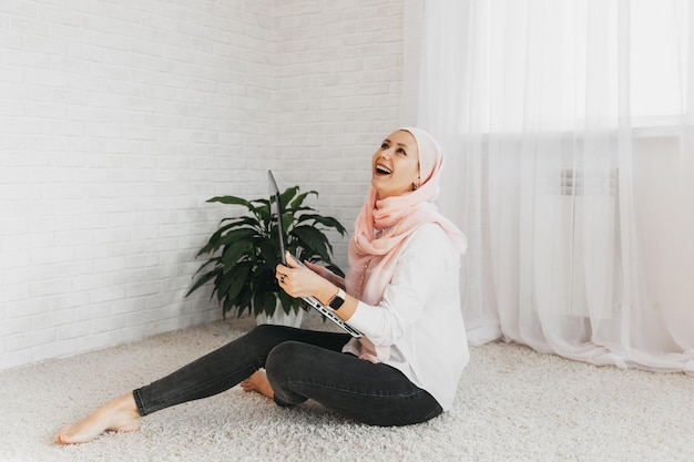 Jonge moderne moslimvrouw in hijab zit op de vloer en werkt thuis op een laptop, online training, werk op afstand.