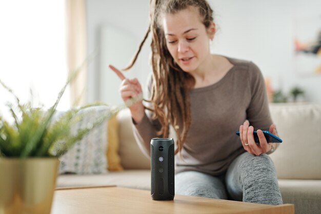 Jonge moderne vrouw met smartphone zittend op de Bank voor slimme spreker en vraag stellen