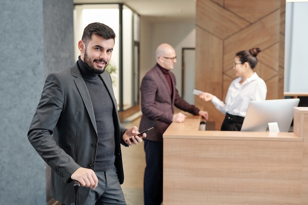 Jonge moderne zakenreiziger met mobiele telefoon die naar u kijkt terwijl u een taxi in de hotellounge belt