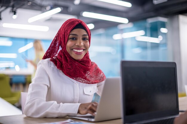 Jonge moderne zwarte moslim zakenvrouw draagt een rode hijab, bezig met laptopcomputer in startbureau. Diversiteit, multiraciaal concept