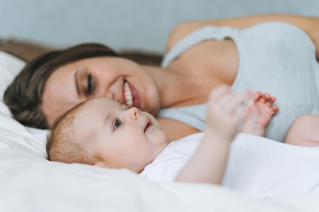 Jonge moeder die plezier heeft met schattige babymeisje op het bed met witte linnen natuurlijke tinten houden van emotie