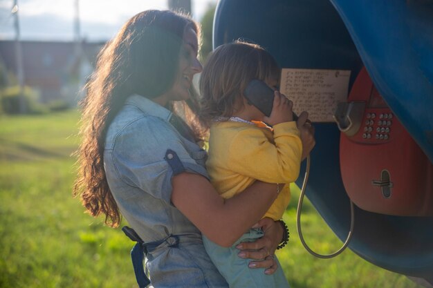 Jonge moeder en baby bellen in de zomer op een vaste straattelefoon in de telefooncel in het dorp