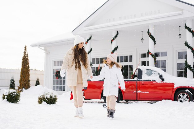 Jonge moeder en dochter in winterkleren houden elkaars hand vast en lopen rond op de winterbinnenplaats van hun huis, versierd voor Kerstmis