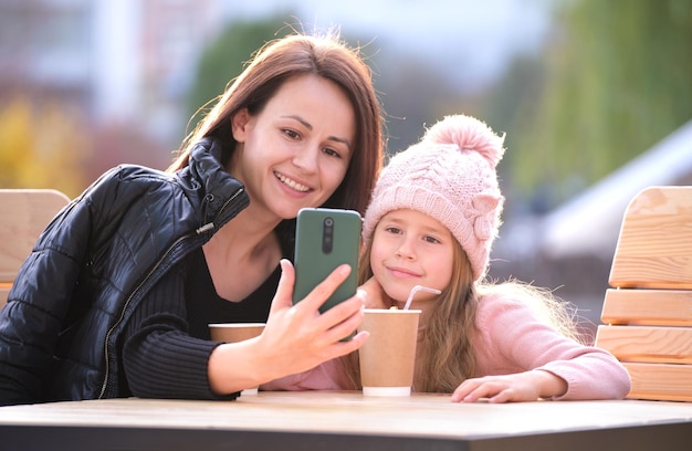 Jonge moeder en haar kind dochter nemen foto met telefoon selfie camera zittend op straat café met warme dranken op zonnige herfstdag Social media aanwezigheid in het dagelijks leven concept