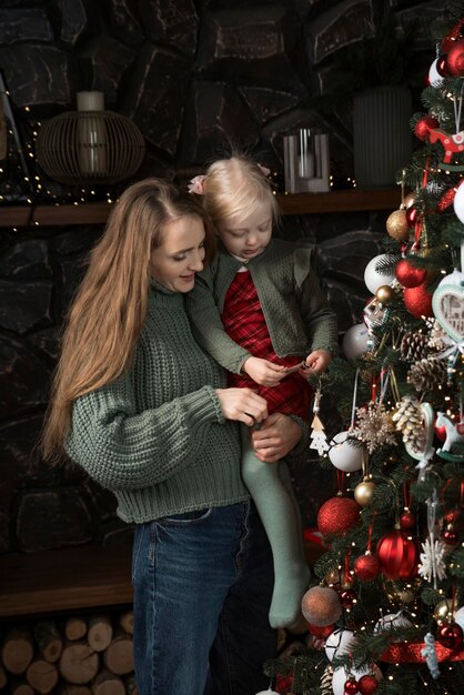 Jonge moeder en kind bij de kerstboom Moeder houdt kleine dochter vast en versiert een prachtige kerstboom Vakanties