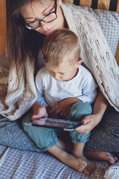 Jonge moeder en zoon met behulp van een tablet onder een gezellige deken in de slaapkamer van hun huis