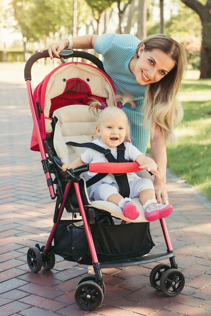 Jonge moeder in gesprek met lachende baby in roze kinderwagen. Ouders lopen buiten met kind in de zomer kinderwagen.