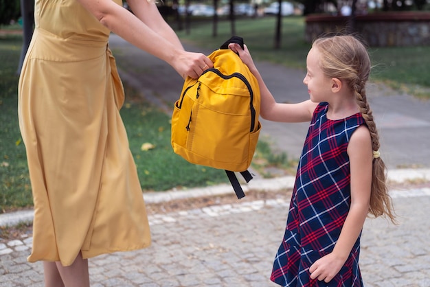 Jonge moeder in lange gele jurk geeft lachende schoolmeisje rugzak voorbereiden op lessen