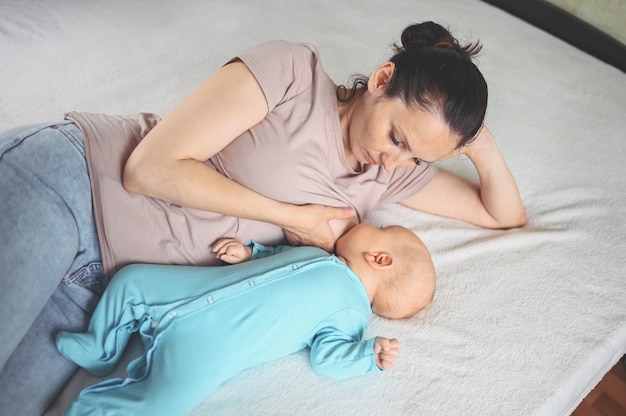Jonge moeder ligt met een pasgeboren schattige baby in blauwe jumpsuit op het bed, knuffelt en geeft hem borstvoeding met moedermelk