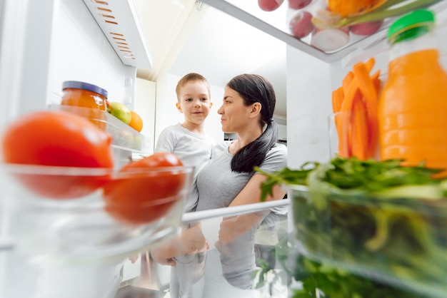 Jonge moeder met baby in de buurt van open koelkast. Gezond eten thuis concept. Groenten en fruit in de koelkast