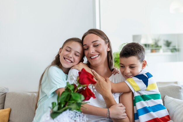 Jonge moeder met een boeket rozen lacht, knuffelt haar zoon en heerful meisje met een kaart feliciteert moeder tijdens vakantieviering in de keuken thuis