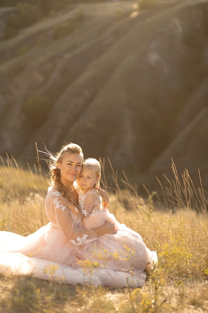Jonge moeder met een dochtertje in roze jurken zit in het veld. Moeder knuffelt dochter, knuffelt haar