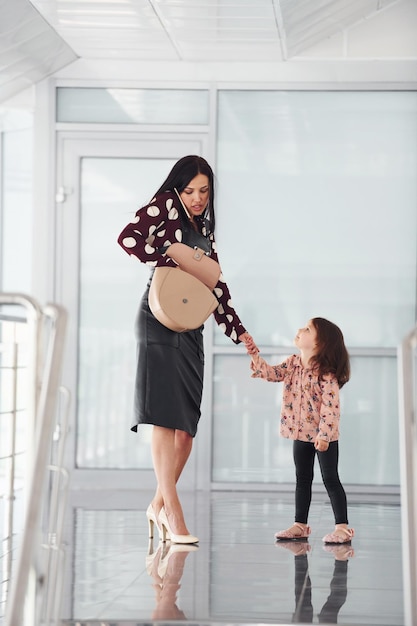 Jonge moeder met haar dochter die samen binnenshuis op kantoor of op de luchthaven loopt. Vakantie hebben